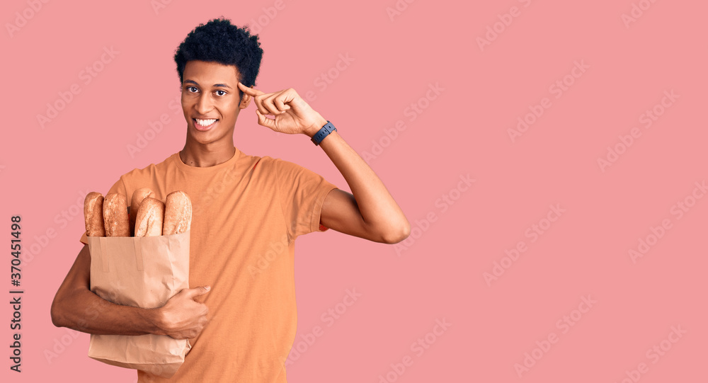 Young african american man holding paper bag with bread smiling pointing to head with one finger, great idea or thought, good memory Young african american man holding paper bag with bread smiling pointing to head with one finger, great idea or thought, good memory