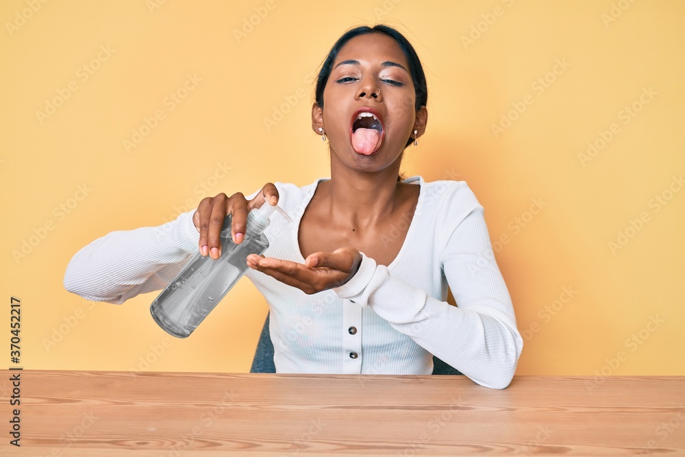 Young indian girl sitting on the table cleaning hand using sanitizer gel sticking tongue out happy with funny expression.
