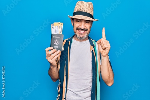 Middle age tourist man on vacation holding united states passport with boarding pass smiling with an idea or question pointing finger with happy face  number one