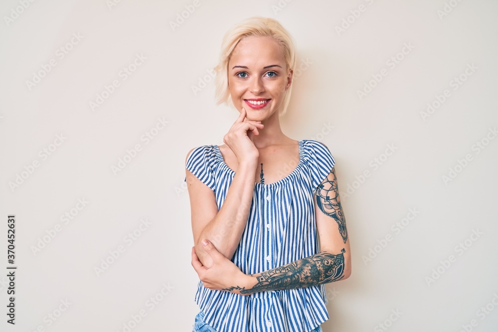 Young blonde woman with tattoo wearing casual clothes smiling looking confident at the camera with crossed arms and hand on chin. thinking positive. Young blonde woman with tattoo wearing casual clothes smiling looking confident at the camera with crossed arms and hand on chin. thinking positive.