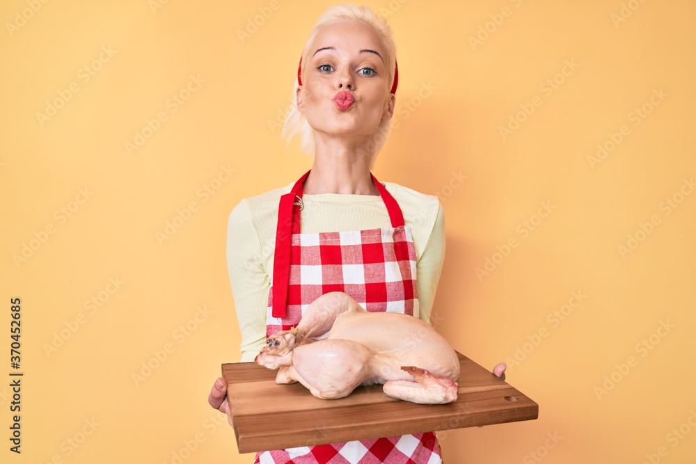Young blonde woman wearing apron cooking chicken looking at the camera blowing a kiss being lovely and sexy. love expression. Young blonde woman wearing apron cooking chicken looking at the camera blowing a kiss being lovely and sexy. love expression.