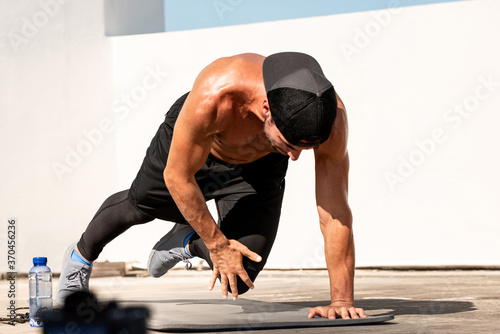 Shirtless athletic man doing mountain climber knee touch exercise outdoors on building rooftop floor in sunlight