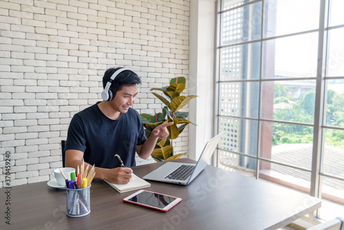 Smiling young asian man wear casual clothing while sitting at big wooden table in modern kitchen, working and video conference meeting from home with computer laptop at home. Social Distancing Concept