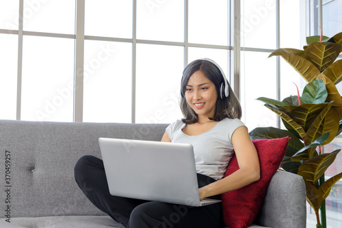 Smiling young asian woman wear casual clothing sitting on cozy couch, working on laptop computer while listen music at home