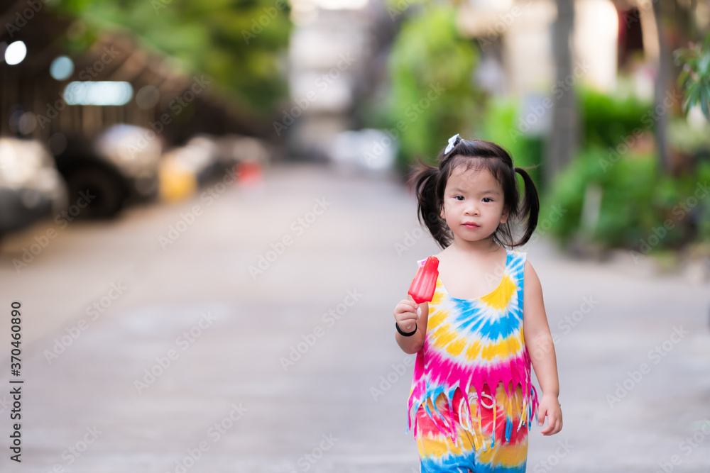 Black-haired little girl standing holding a red popsicle and smiling sweetly during summer or spring. Cute child wear colorful clothes. Children aged 3 - 4 years old. Black-haired little girl standing holding a red popsicle and smiling sweetly during summer or spring. Cute child wear colorful clothes. Children aged 3 - 4 years old.