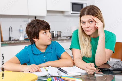 Annoyed mother helping son with homework sitting nearby at kitchen table