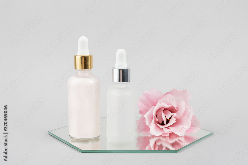 Two glass bottles with serum, essential oil or other cosmetics product and beautiful pink flowers on mirror on grey background. Natural Organic Spa Cosmetic Beauty concept Front view Two glass bottles with serum, essential oil or other cosmetics product and beautiful pink flowers on mirror on grey background. Natural Organic Spa Cosmetic Beauty concept Front view