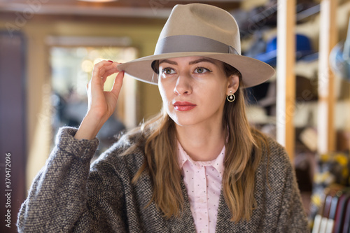 Portrait of elegant young woman trying on new hat during shopping in fashion showroom