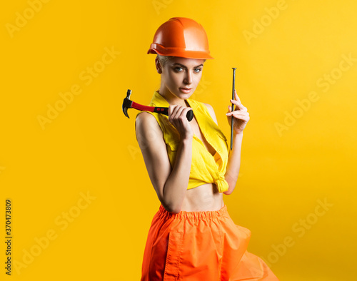 Sexy woman in helmet to use hammer. Young woman hammering nail at workshop on yellow background isolated. Female model working on building. Everything can be fixed concept. © Volodymyr