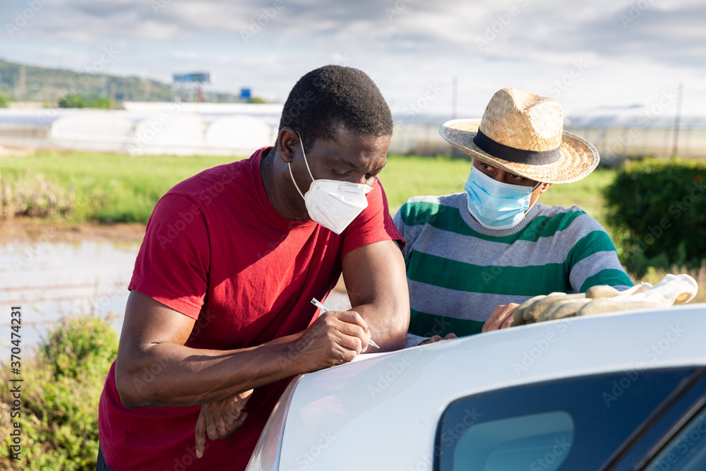 Farmer and worker in medical masks signing papers standing near car on farm field. Pandemic prevention and social distancing concept