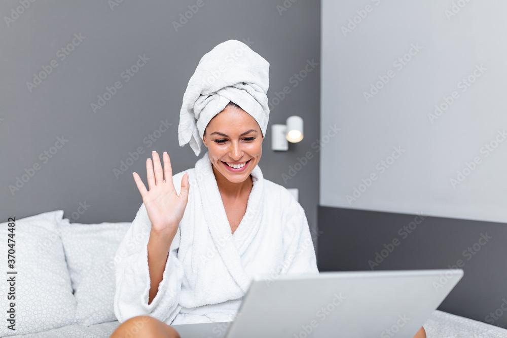 Beautiful young woman in white bathrobe in bedroom at home taking to her friends via video call . Smiling young woman lying on bed in bathrobe relaxing on the bed after bath and looking at her laptop Beautiful young woman in white bathrobe in bedroom at home taking to her friends via video call . Smiling young woman lying on bed in bathrobe relaxing on the bed after bath and looking at her laptop