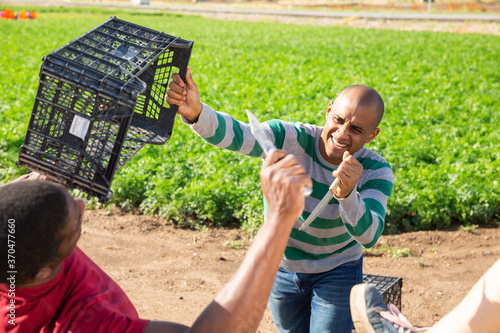 Aggressive hispanic farm worker with knife and plastic box in hands fighting with his african workmate on field photo