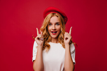 Ecstatic white girl in beret posing with amazement. Elegant caucasian female model in t-shirt standing on red background.