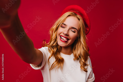 Wonderful white girl in beret making selfie with eyes closed and smiling. Portrait of adorable blonde female model with red lips.