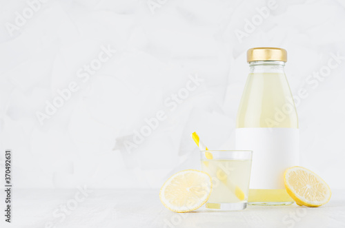 Bright yellow lemon juice in glass bottle mock up with blank label, straw, wine glass, fruit slice on white wood table in light interior, template for packaging, advertising, design, branding product.