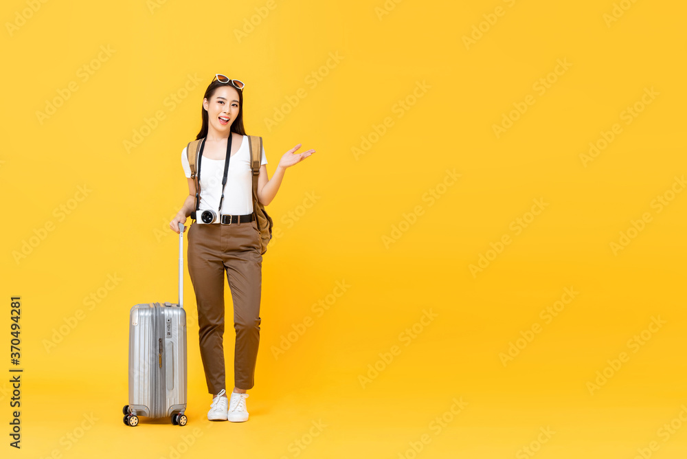 Full length portrait of smiling cute young Asian woman tourist with trolley bag opening hand to copy space aside isolated on yellow studio background Full length portrait of smiling cute young Asian woman tourist with trolley bag opening hand to copy space aside isolated on yellow studio background