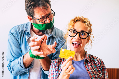 Crazy and cheerful nice beautiful adult woman take bank credit card ready to buy and blocked angry man husband in background - funny economy concept scene at home with people