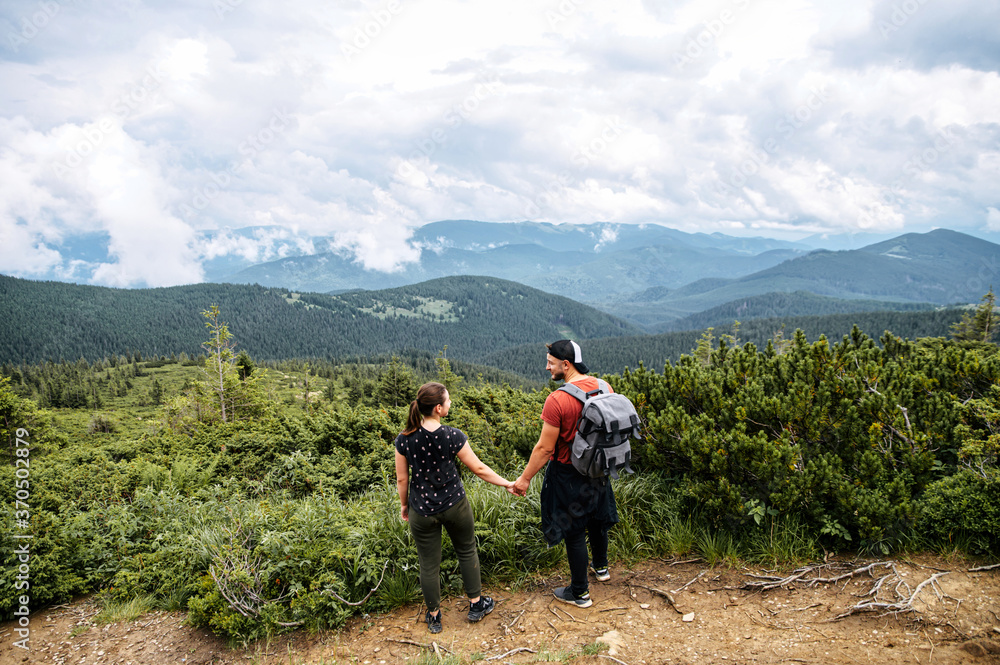 Back view of a guy and a girl hikers stand and holding hands with a spectacular view. Couple in love hike in a mountains