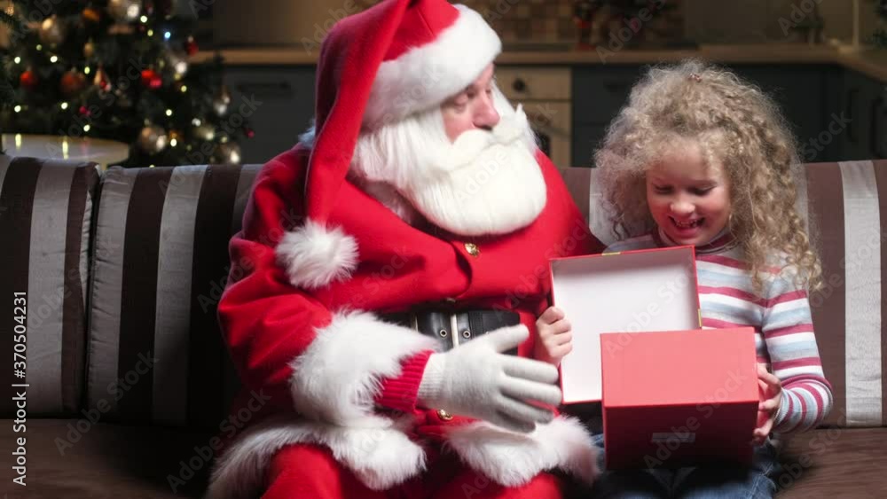 Santa Claus in a red suit with a beard and a cap gives a gift for Christmas to a curly-haired happy girl. The child opens the box and is happy with the gift. waiting for wonder at Christmas Santa Claus in a red suit with a beard and a cap gives a gift for Christmas to a curly-haired happy girl. The child opens the box and is happy with the gift. waiting for wonder at Christmas