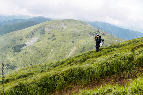 Photographer is taking a photo in the mountains. A hiker guy with a backpack stands on footpath with a professional camera with a scenic view on the background