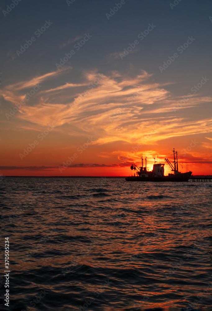 Beautiful orange sunset on sea water. Transport ship on the dock in the evening on seacoast. Shipping logistic by sea. Amazing marine landscape with colorful cloudy sky.