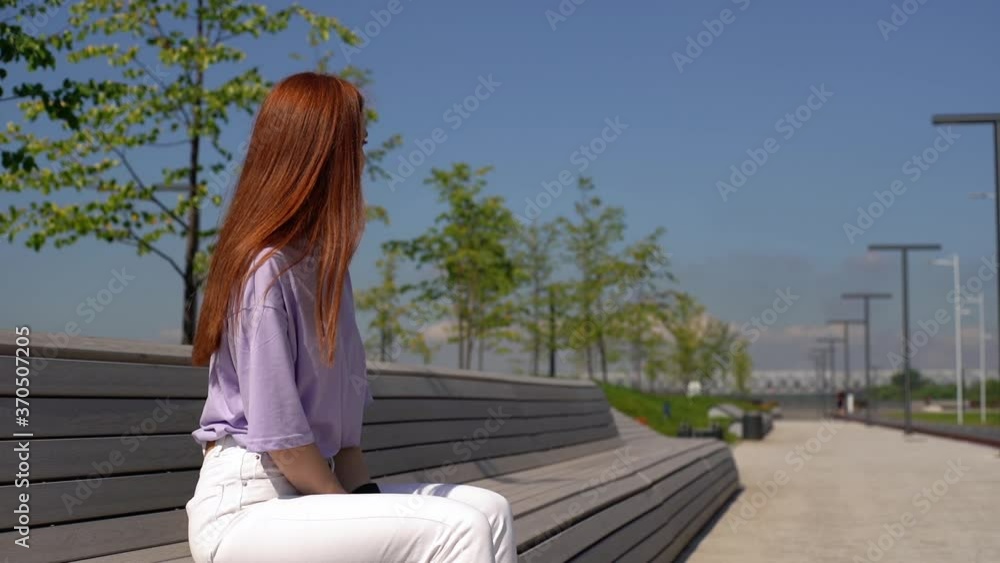 Sad pretty red-haired girl wearing fashion casual clothes sitting on bench in city park on bright sunny day. Upset attractive young woman contemplating outdoors. Shooting in slow motion. Sad pretty red-haired girl wearing fashion casual clothes sitting on bench in city park on bright sunny day. Upset attractive young woman contemplating outdoors. Shooting in slow motion.