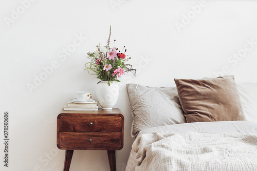 Cup of coffee and books on retro wooden bedside table. Rustic white ceramic vase with bouquet of pink cocmos and zinnia flowers. Beige linen and velvet pillows in bed. Scandinavian interior, bedroom. photo