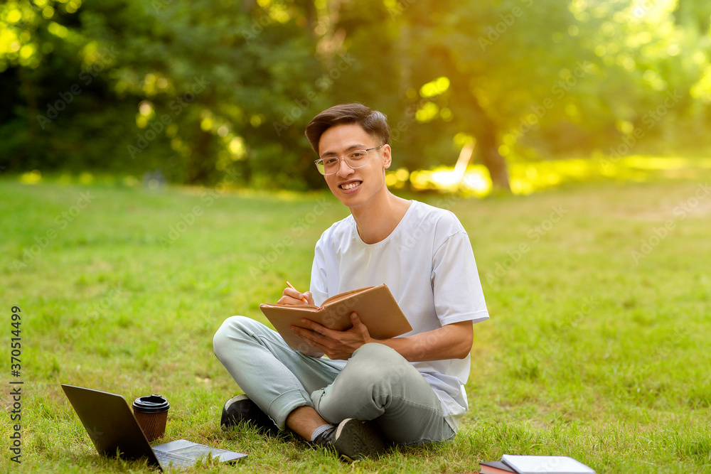 Teen Asian Guy Studying With Book Outdoors In Park And Taking Notes Teen Asian Guy Studying With Book Outdoors In Park And Taking Notes
