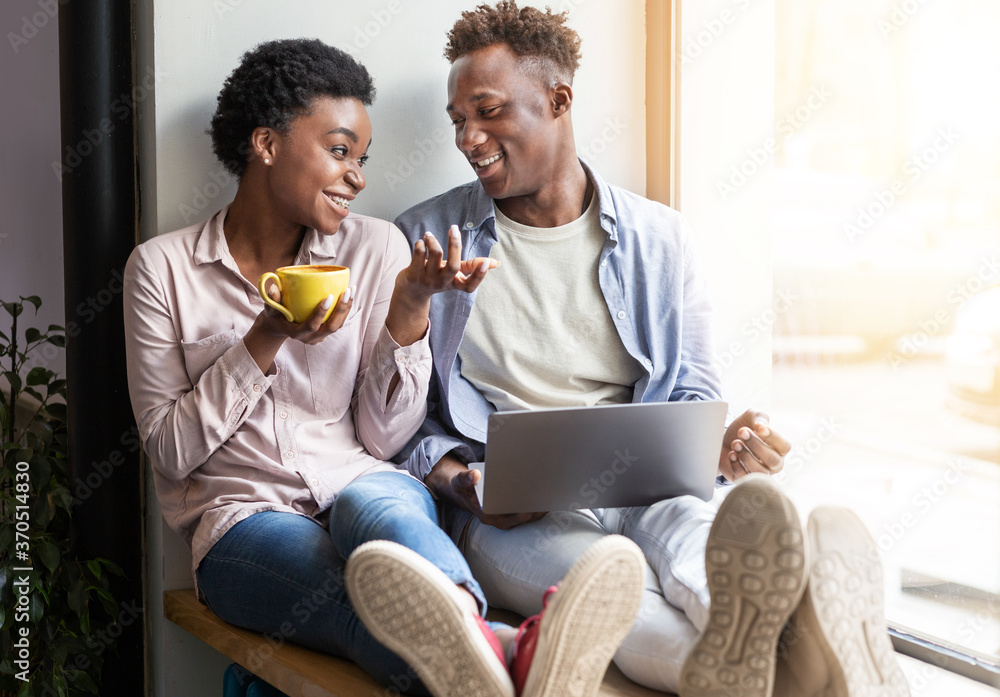 Joyful African American couple with laptop and coffee sitting on windowsill at cafe