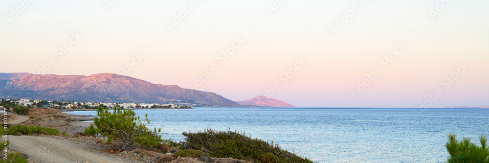 seascape at dusk. a dirt road, a city in the distance and mountains with a gentle pink sunset and a beautiful blue sea. banner
