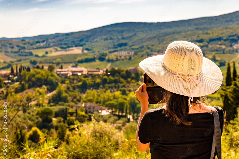 A woman, wearing a big sun bonnet, is taking photos with her mobile phone of the beautiful countryside of the famous medieval town San Gimignano in Tuscany. The picture focuses on the woman.