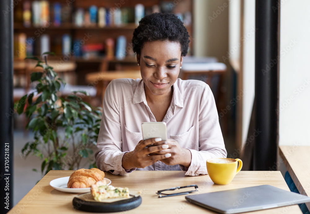 Young black woman using mobile phone for online communication at lunchtime in coffee shop Young black woman using mobile phone for online communication at lunchtime in coffee shop