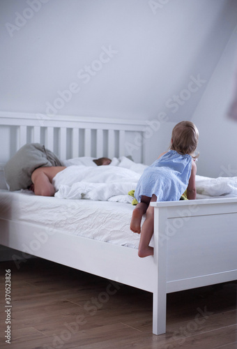 A little girl in a blue dress climbs onto a bed in her parents' bedroom. Minimalist interior, white bed linen. Dad sleeps