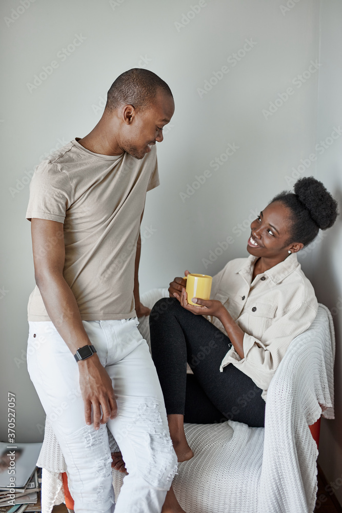good-looking afro couple have rest at home, woman sits with yellow mug in hands, man talk with her. isolated in light room good-looking afro couple have rest at home, woman sits with yellow mug in hands, man talk with her. isolated in light room