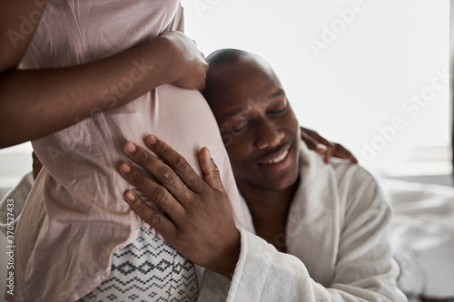 happy young african man is listening to the belly of his pregnant wife, couple in domestic wear at home
