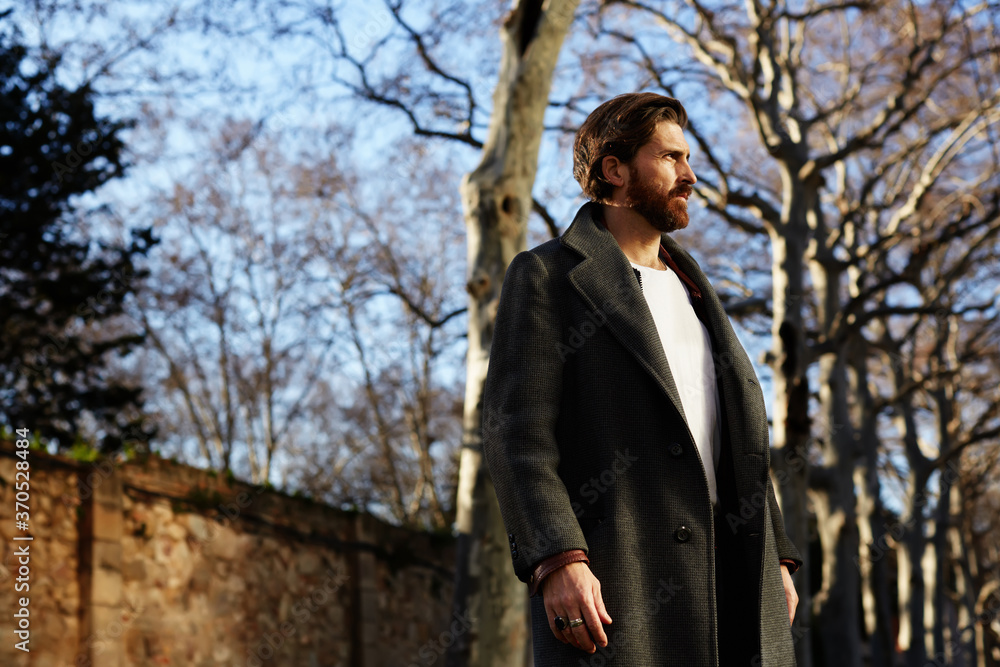 Portrait of fashionable well dressed man with beard posing outdoors looking away, confident and focused mature man in coat standing outside at sunny evening, elegant fashion model Portrait of fashionable well dressed man with beard posing outdoors looking away, confident and focused mature man in coat standing outside at sunny evening, elegant fashion model