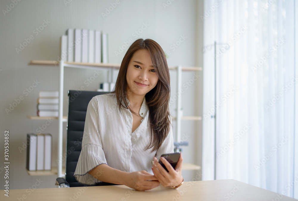 Young beautiful Asian business woman sitting at her working desk and using smartphone with relaxing expression. Young beautiful Asian business woman sitting at her working desk and using smartphone with relaxing expression.