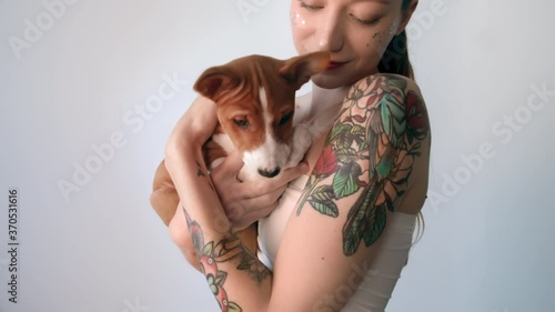 Closeup of beautiful tattooed young woman with makeup holding cute basenji puppy dog on hands. Tender and love pets animals happy together