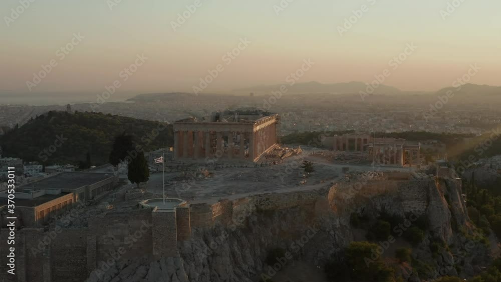 Aerial Flight towards Acropolis of Athens with Greek Flag Waving in Beautiful Golden Hour Sunset light with Ocean in the distance