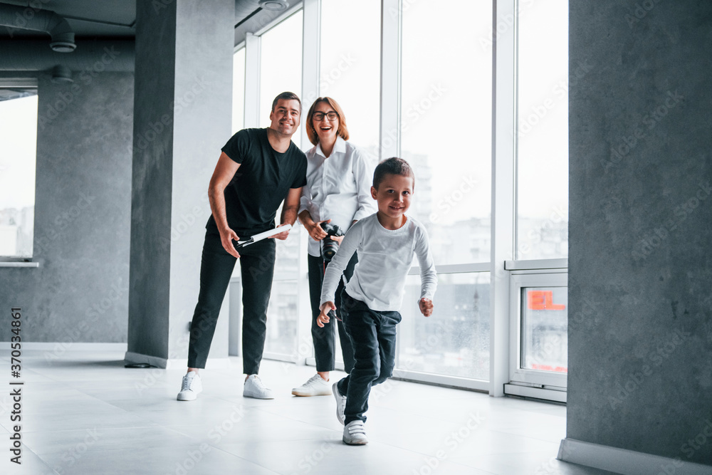 Woman in formal clothes and with camera in hand standing inside of empty room with man and little boy that running and having fun Woman in formal clothes and with camera in hand standing inside of empty room with man and little boy that running and having fun