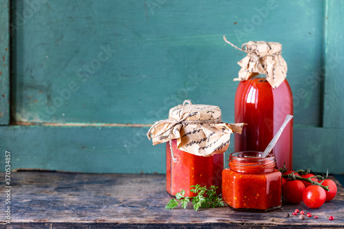 Homemade tomato ketchup made from ripe red tomatoes in glass jars with ingredients on an old wooden table. Copy space