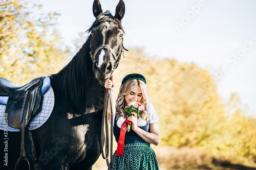 retty blonde in traditional dress walking with big black horse outdoors