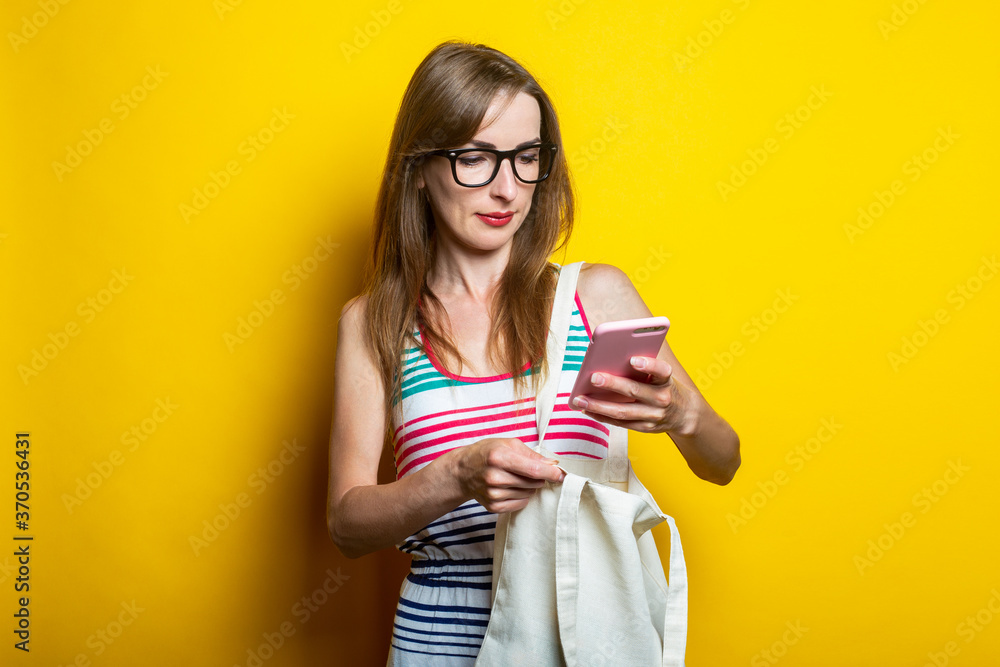 Beautiful smiling young woman in glasses with a linen bag holding a phone on a yellow background. Beautiful smiling young woman in glasses with a linen bag holding a phone on a yellow background.