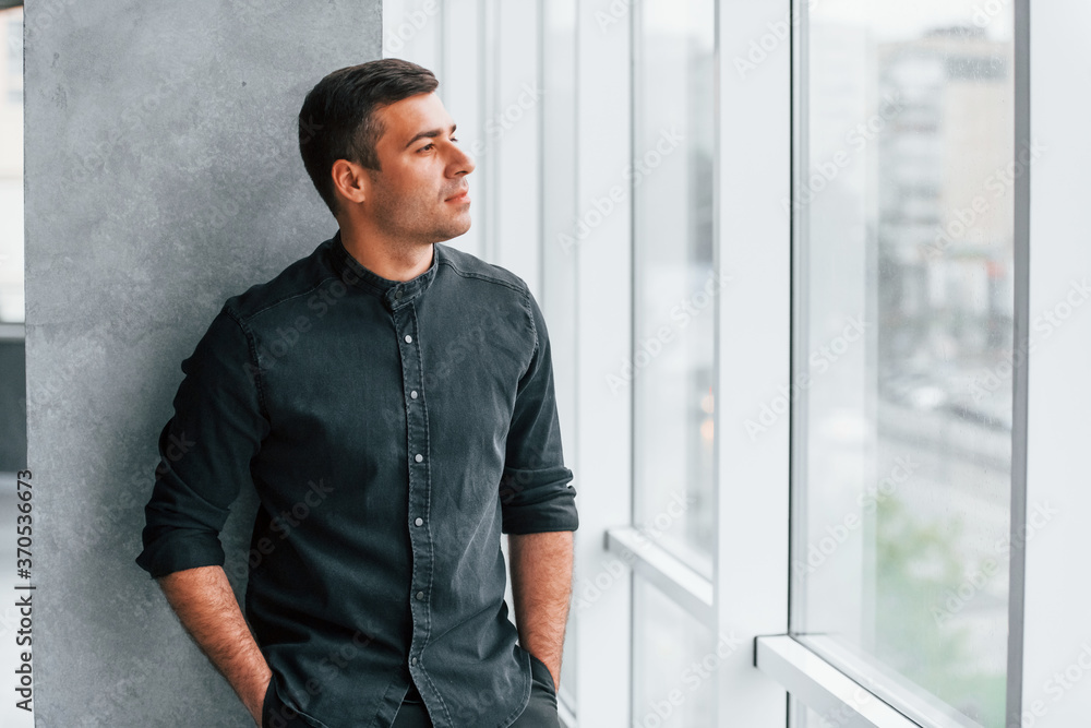 Elegant man in black shirt leaning on the wall indoors and looking through the big window Elegant man in black shirt leaning on the wall indoors and looking through the big window