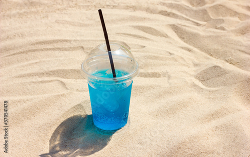 Blue drink cocktail with ice in a plastic glass with a straw on the beautiful sand, a cold refreshing drink in a hot gin, the theme of travel and travel by the sea