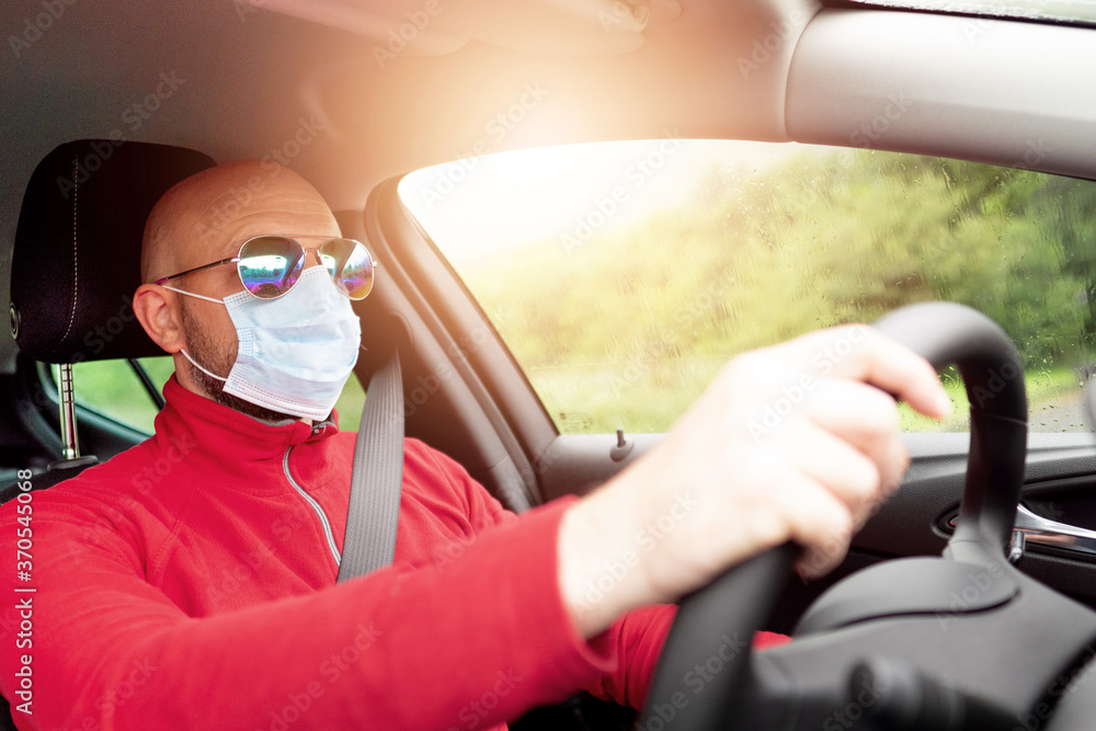 Bald man in red shirt, reflective glasses and blue mask driving a car on a sunny day. COVID 19 pandemic prevention concept. Bald man in red shirt, reflective glasses and blue mask driving a car on a sunny day. COVID 19 pandemic prevention concept.