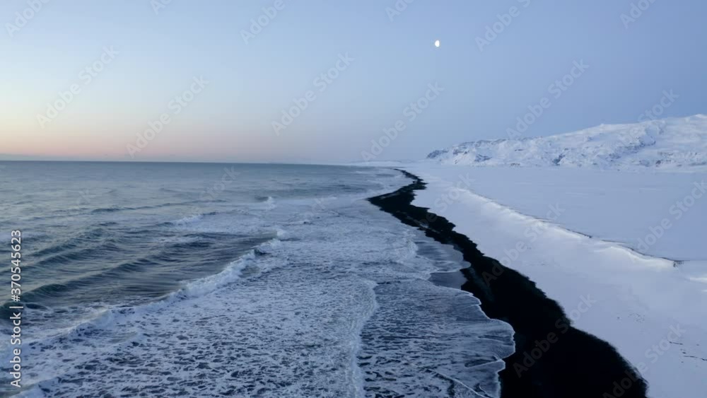 AERIAL: Flying over Black Beach with white arctic snow mountains in background in Iceland in Winter Snow, Ice, Waves, Water AERIAL: Flying over Black Beach with white arctic snow mountains in background in Iceland in Winter Snow, Ice, Waves, Water