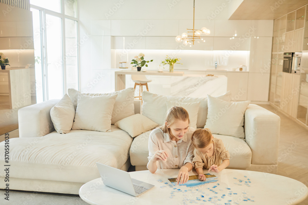 Warm-toned wide angle portrait of two sisters solving puzzle together while enjoying time at home indoors in minimal white interior, copy space