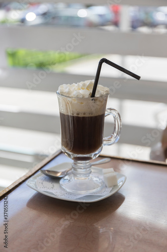 There is a clear glass of coffee on the wooden table. In the background - napkins.Pipe for drinks.