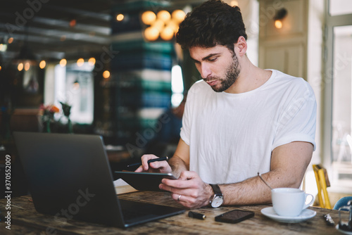 Handsome director of coffee shop concentrated working on touch pad searching new ideas for own business using 4G wireless internet connection and modern gadgets sitting at the table with cup of coffee